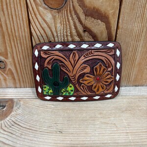K&ouml;nnte beinhalten: Rechteckige, braune Lederg&uuml;rtelschnalle mit detailliertem, geschnitztem Design. Das Design zeigt einen gr&uuml;nen Kaktus, eine orangefarbene Blume mit roter Mitte und wirbelnde Blumenmuster. Die R&auml;nder sind mit wei&szlig;en Rauten verziert.