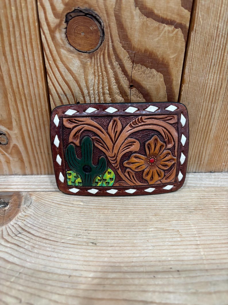 K&ouml;nnte beinhalten: Rechteckige, braune Lederg&uuml;rtelschnalle mit detailliertem, geschnitztem Design. Das Design zeigt einen gr&uuml;nen Kaktus, eine orangefarbene Blume mit roter Mitte und wirbelnde Blumenmuster. Die R&auml;nder sind mit wei&szlig;en Rauten verziert.