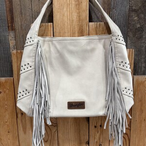 K&ouml;nnte beinhalten: Cremefarbene Schultertasche mit Fransen und Nieten. Die Tasche hat eine rechteckige Form mit einem Griff und einem kleinen braunen Lederaufn&auml;her mit dem Wort "Wrangler". Die Tasche ist aus weichem Material.