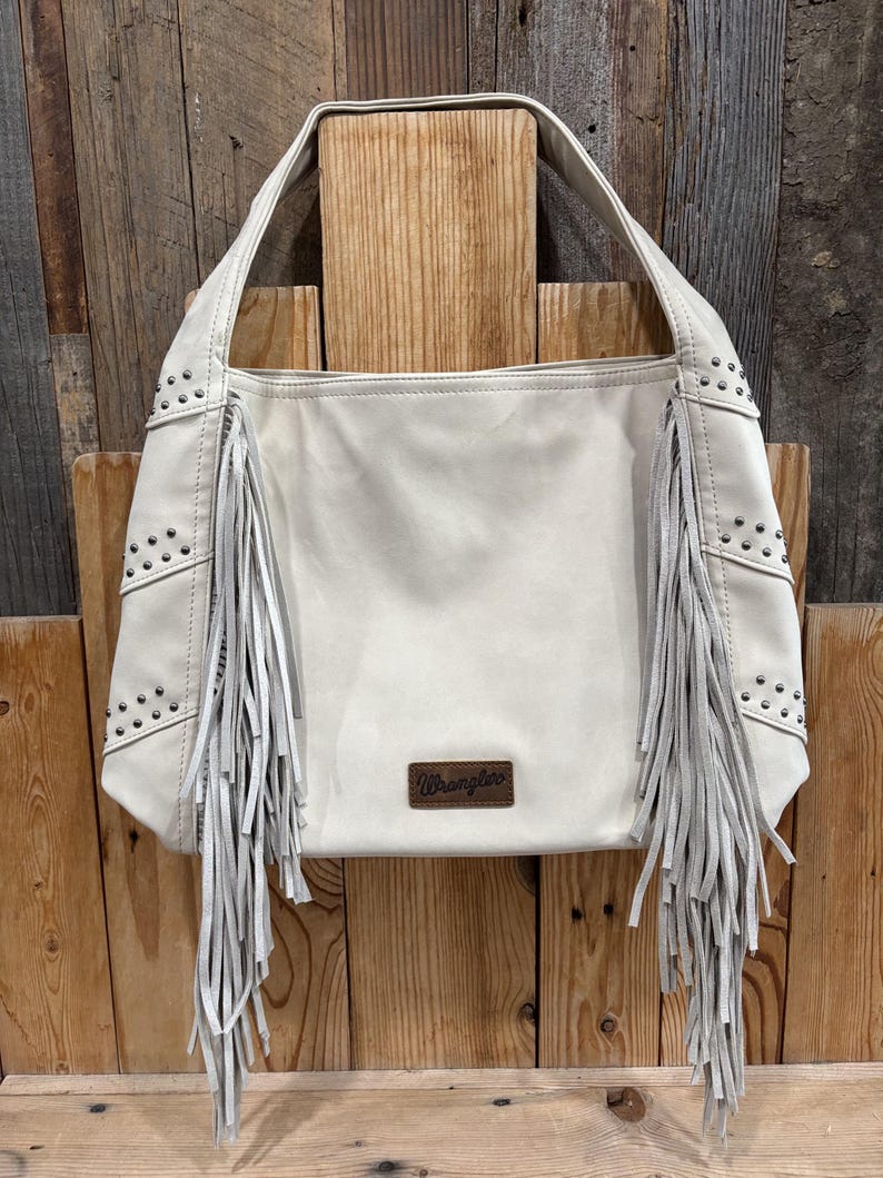 K&ouml;nnte beinhalten: Cremefarbene Schultertasche mit Fransen und Nieten. Die Tasche hat eine rechteckige Form mit einem Griff und einem kleinen braunen Lederaufn&auml;her mit dem Wort "Wrangler". Die Tasche ist aus weichem Material.