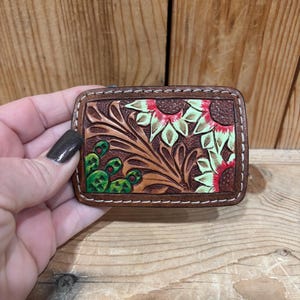 K&ouml;nnte beinhalten: Eine rechteckige, braune Ledergeldb&ouml;rse mit einem Blumendesign mit Sonnenblumen und Kakteen. Das Design ist gepr&auml;gt und mit roten, gr&uuml;nen und hellgr&uuml;nen Akzenten bemalt. Die Kanten sind mit wei&szlig;em Faden gen&auml;ht.