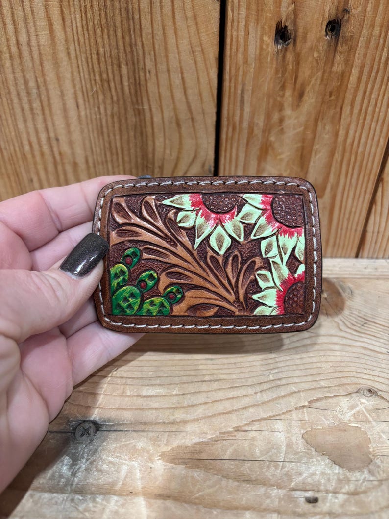 K&ouml;nnte beinhalten: Eine rechteckige, braune Ledergeldb&ouml;rse mit einem Blumendesign mit Sonnenblumen und Kakteen. Das Design ist gepr&auml;gt und mit roten, gr&uuml;nen und hellgr&uuml;nen Akzenten bemalt. Die Kanten sind mit wei&szlig;em Faden gen&auml;ht.