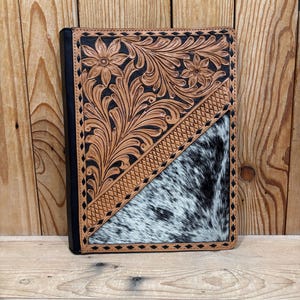 Op de afbeelding: Een leren notitieboek met een zwarte rug en een decoratieve omslag. De omslag heeft een bloemmotief in bruin leer, een diagonale sectie van koeienhuid en een zwart-wit patroon.