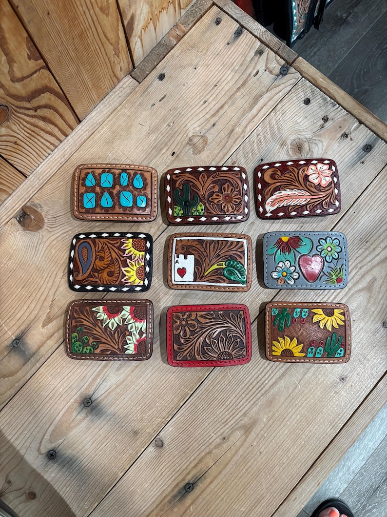 K&ouml;nnte beinhalten: Neun rechteckige G&uuml;rtelschnallen aus Leder mit verschiedenen Designs. Die Schnallen weisen florale, Kaktus- und geometrische Muster auf, mit Farben wie T&uuml;rkis, Rot, Gelb und Wei&szlig;. Die Schnallen werden auf einer Holzoberfl&auml;che pr&auml;sentiert.