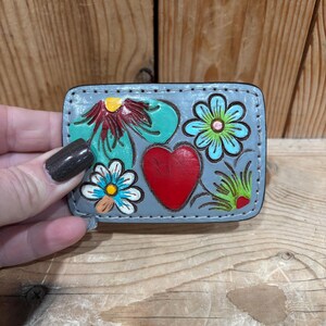 K&ouml;nnte beinhalten: Kleines, rechteckiges, graues Lederst&uuml;ck mit handgemaltem Blumen- und Herzdesign. Das Design umfasst ein rotes Herz, t&uuml;rkisfarbene Blumen und gelbe und blaue Akzente. Der Artikel wird vor einem h&ouml;lzernen Hintergrund gehalten.