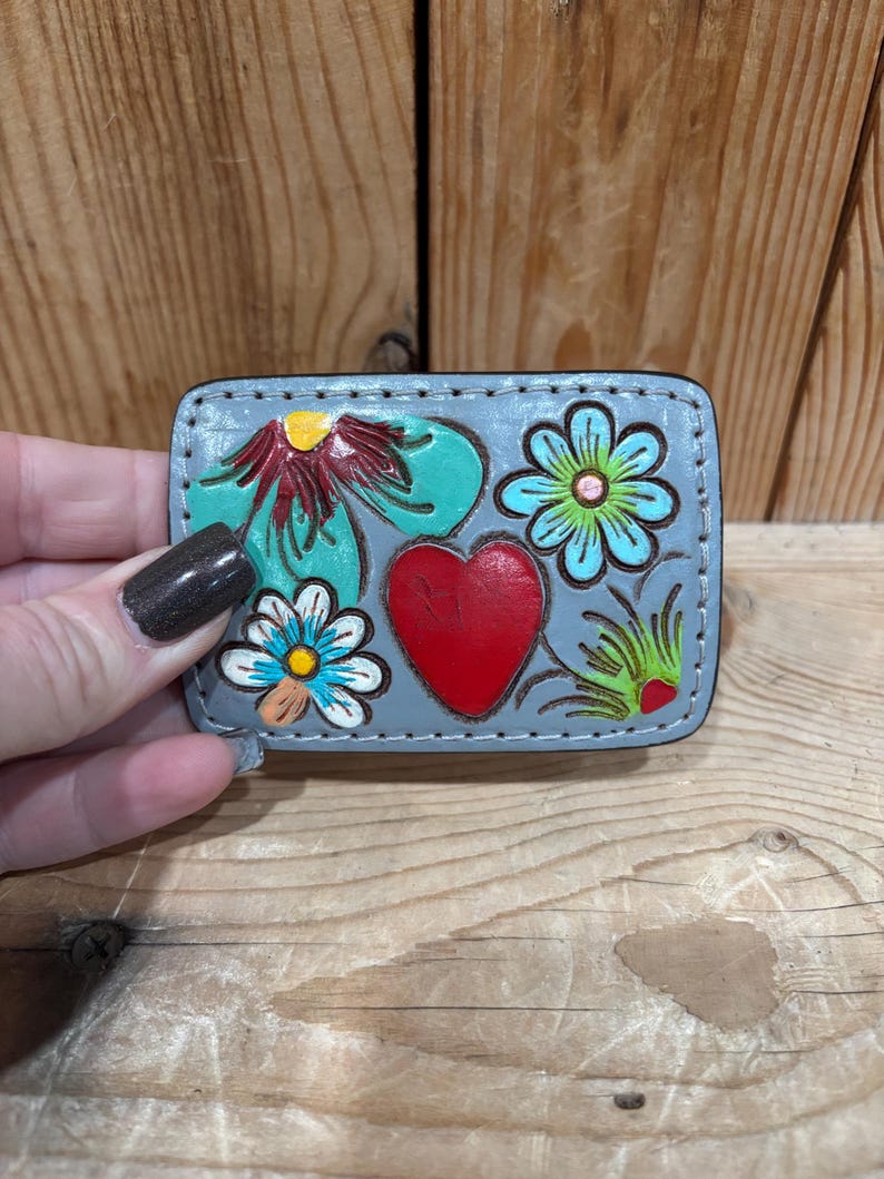 K&ouml;nnte beinhalten: Kleines, rechteckiges, graues Lederst&uuml;ck mit handgemaltem Blumen- und Herzdesign. Das Design umfasst ein rotes Herz, t&uuml;rkisfarbene Blumen und gelbe und blaue Akzente. Der Artikel wird vor einem h&ouml;lzernen Hintergrund gehalten.