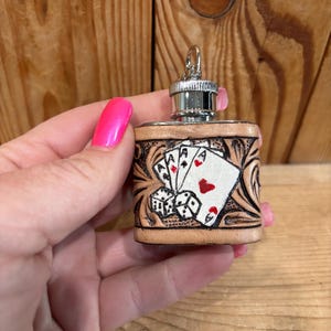 Op de afbeelding: Kleine, bruine leren flacon met een zilveren dop. De flacon is versierd met een speelkaartenontwerp met azen en dobbelstenen. Het leer heeft een gesneden bloemenpatroon rond het kaartontwerp.
