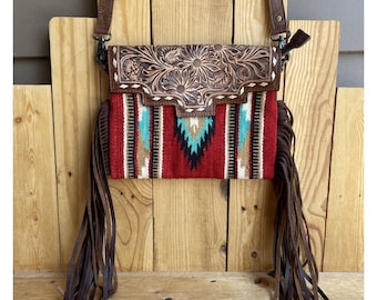 May include: A crossbody bag with a southwestern design. The bag features a red, turquoise, and white patterned fabric body with brown leather accents. The top flap is embossed with a floral design. Long brown fringe hangs from the bottom.