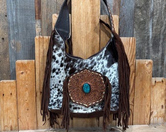 May include: A hobo-style shoulder bag featuring a black and white cowhide pattern with long brown leather fringe accents. The front displays a hexagonal brown leather patch with intricate floral tooling and a central oval turquoise stone.