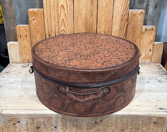 May include: A round, brown leather hat box featuring an intricate, embossed floral and leaf pattern across the lid and sides. The container includes a sturdy leather handle attached to the front and metal hardware rings on the sides.