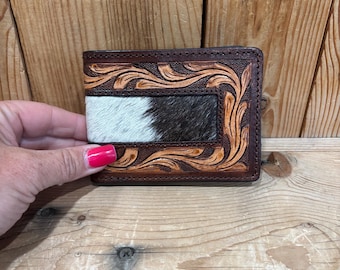 May include: A bifold wallet featuring a central panel of black and white cowhide hair-on leather surrounded by a dark brown leather border with an intricate, hand-tooled floral scroll pattern and visible stitching along the edges.