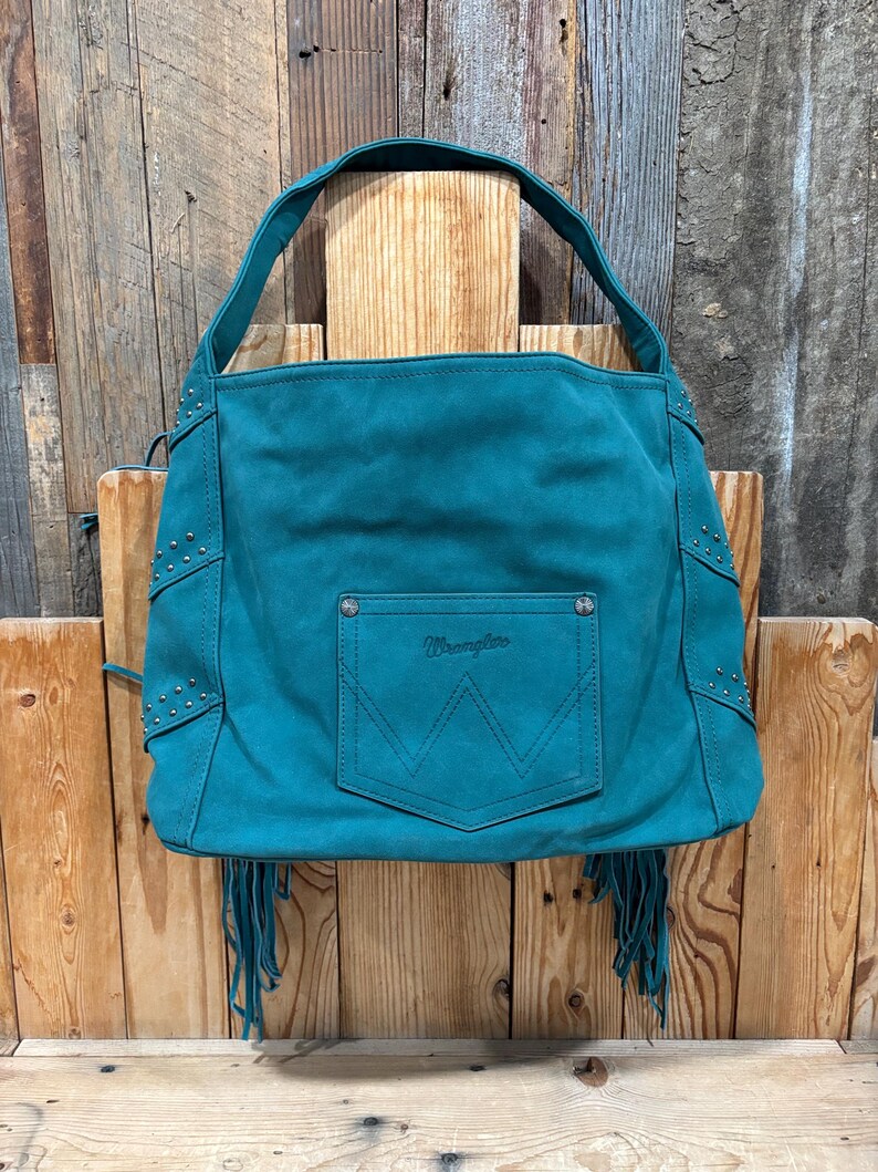 K&ouml;nnte beinhalten: T&uuml;rkisfarbene Wildleder-Umh&auml;ngetasche mit oberem Griff und Vordertasche. Die Tasche hat silberne Nieten und Fransen. Das Wrangler-Logo ist auf der Tasche zu sehen.
