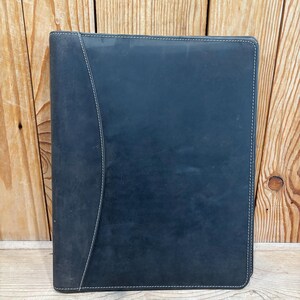 Op de afbeelding: Een donkerblauwe leren portfolio met witte stiksels langs de randen en een gebogen accent. De portfolio is gesloten en lijkt gemaakt van een textuurrijk materiaal. De achtergrond is een houten oppervlak.