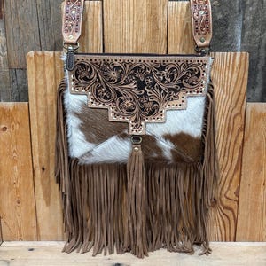 May include: A brown and white cowhide crossbody bag with a decorative floral tooled leather flap. The bag features long brown fringe, a tassel, and leather straps with decorative stitching. The bag is designed with a Western theme.
