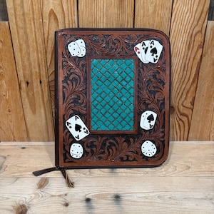 Op de afbeelding: Een bruine leren kaarthouder met een centraal turquoise paneel en decoratieve bloemmotieven. De houder heeft speelkaarten en dobbelsteenontwerpen. Een rits en een leren trekker zijn zichtbaar.