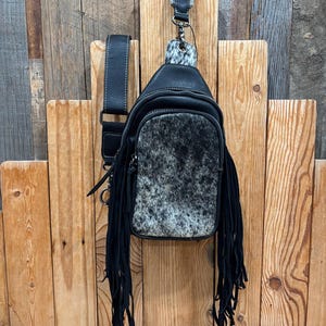 May include: A black crossbody sling bag featuring a front panel of speckled gray and black cowhide hair-on leather. The bag includes long black fringe accents on both sides, a black adjustable shoulder strap, and a matching cowhide leather top tab.