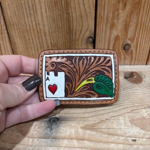 K&ouml;nnte beinhalten: Rechteckige, handgefertigte G&uuml;rtelschnalle aus Leder mit hellbraunem Rand. Das Design zeigt das Herz-Ass, ein Blumenmuster und ein gr&uuml;nes Blatt mit gelben Akzenten. Die Schnalle wird von einer Person gehalten.