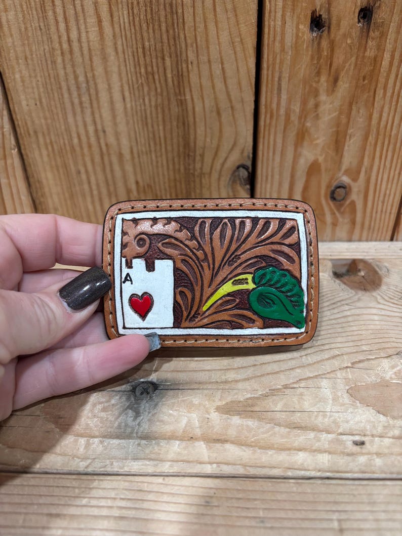 K&ouml;nnte beinhalten: Rechteckige, handgefertigte G&uuml;rtelschnalle aus Leder mit hellbraunem Rand. Das Design zeigt das Herz-Ass, ein Blumenmuster und ein gr&uuml;nes Blatt mit gelben Akzenten. Die Schnalle wird von einer Person gehalten.