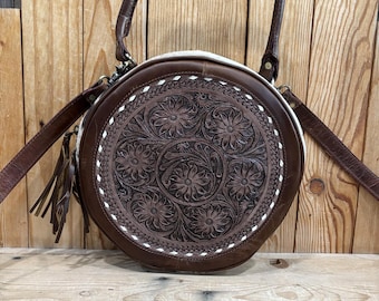 May include: A round, brown leather handbag with a floral embossed design. The bag features a white stitched border, a top handle, and a detachable shoulder strap. A tassel hangs from the side. The bag is set against a wooden background.