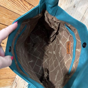 K&ouml;nnte beinhalten: T&uuml;rkisfarbene Tragetasche mit braunem Innenraum, der ein Seilmuster und das "Wrangler"-Logo aufweist. Die Tasche hat Fransen und ein Lederetikett. Die Tasche ist offen und zeigt das Innenfutter.