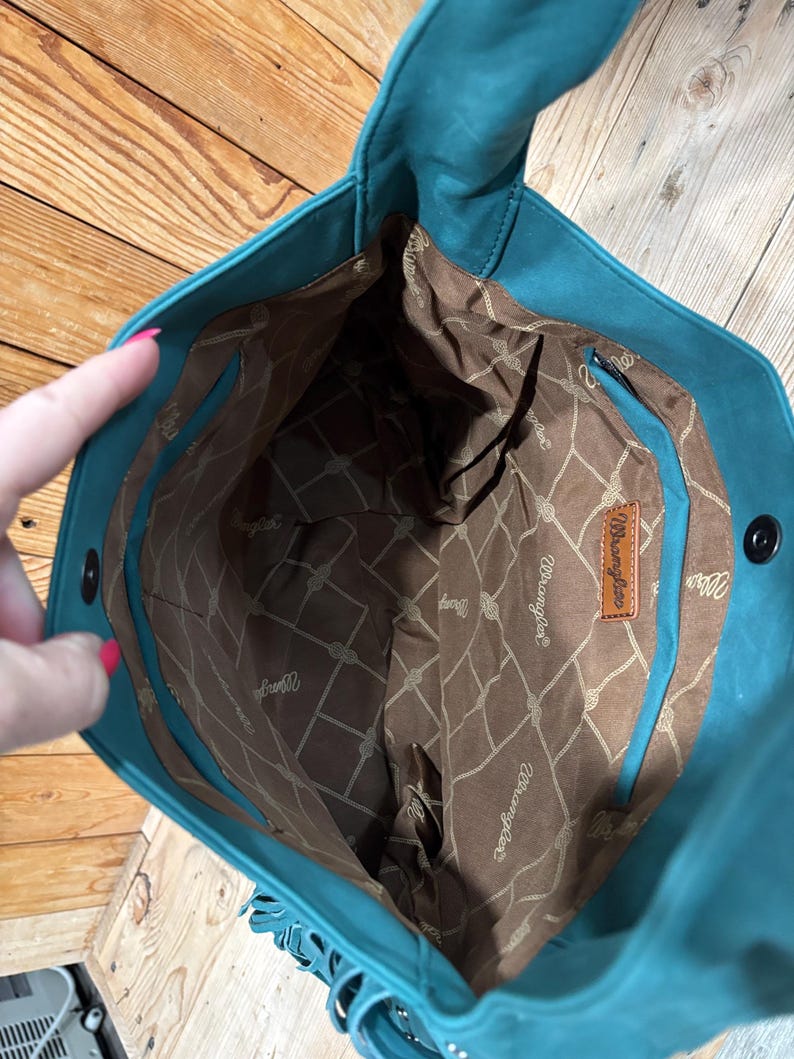 K&ouml;nnte beinhalten: T&uuml;rkisfarbene Tragetasche mit braunem Innenraum, der ein Seilmuster und das "Wrangler"-Logo aufweist. Die Tasche hat Fransen und ein Lederetikett. Die Tasche ist offen und zeigt das Innenfutter.