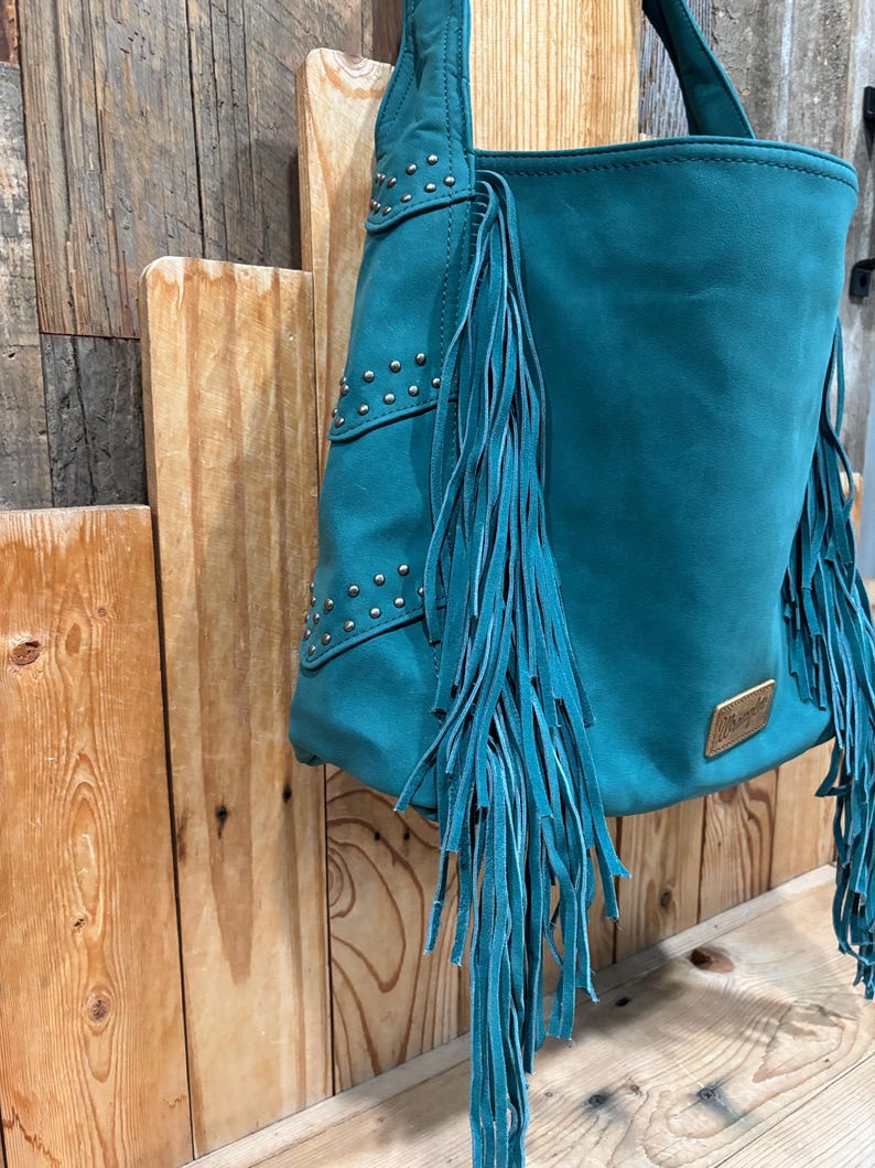 K&ouml;nnte beinhalten: T&uuml;rkisfarbene Ledertasche mit Fransen und silbernen Nieten. Die Tasche hat eine rechteckige Form mit einem Griff und einem kleinen goldfarbenen Etikett. Die Tasche h&auml;ngt an einer Holzoberfl&auml;che.