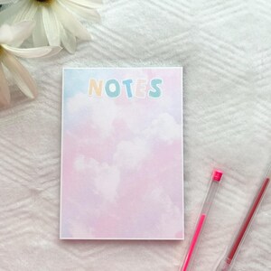 May include: A pink and blue notepad with a watercolor cloud design and the word "NOTES" in colorful letters. Two pink highlighter pens are lying on a white surface next to the notepad.
