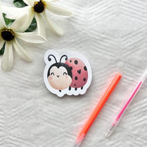 May include: A cute ladybug sticker with a smiling face and a red and black spotted shell. The sticker is on a white background with two white flowers and two pens. The ladybug sticker is a fun and whimsical design.