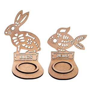 May include: Two wooden candle holders shaped like a fish and a rabbit. The fish has the word "NEMO" on the base and the rabbit has the word "BUGS" on the base. Both are decorated with a Day of the Dead style design.
