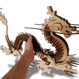 May include: A wooden 3D puzzle of a dragon with intricate details. The dragon is curled up with its head raised and its tail curled around its body. The puzzle is made of laser-cut wood and is assembled by hand.