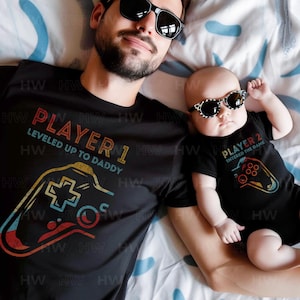 May include: A black t-shirt with the text "Player 1 Leveled Up To Daddy" and a retro-style video game controller graphic. A black baby onesie with the text "Player 2 Entered The Game" and a retro-style video game controller graphic.