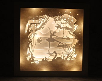 Könnte beinhalten: Eine weiße Leuchtbox mit einem ausgeschnittenen Papierdesign einer Ballerina, die in einem Wald mit Schwänen tanzt. Die Box ist mit warmweißen LED-Lichtern beleuchtet.