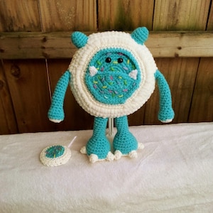 May include: A handmade crochet monster toy in white and turquoise. The round body has a turquoise center with black eyes and colorful sprinkles. It has turquoise arms, legs, and small ears. A matching coaster is next to the toy.