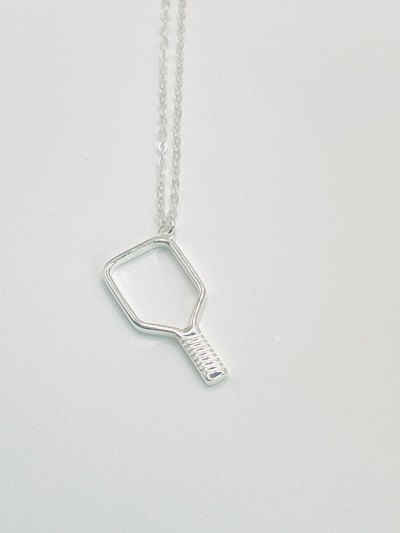 Peut inclure: Un collier en argent avec un pendentif en forme de raquette de pickleball.