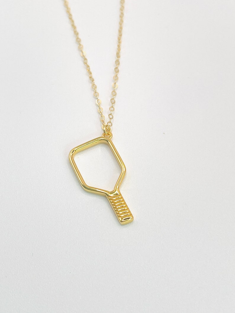 Peut inclure: Un collier en or avec un pendentif en forme de raquette de pickleball.