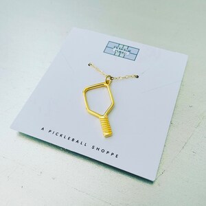 Peut inclure: Un collier en or avec un pendentif en forme de raquette de pickleball. Le pendentif est un simple contour d'une raquette avec un manche textur&eacute;. Le collier est pr&eacute;sent&eacute; sur une carte blanche avec le texte "A Pickleball Shoppe".