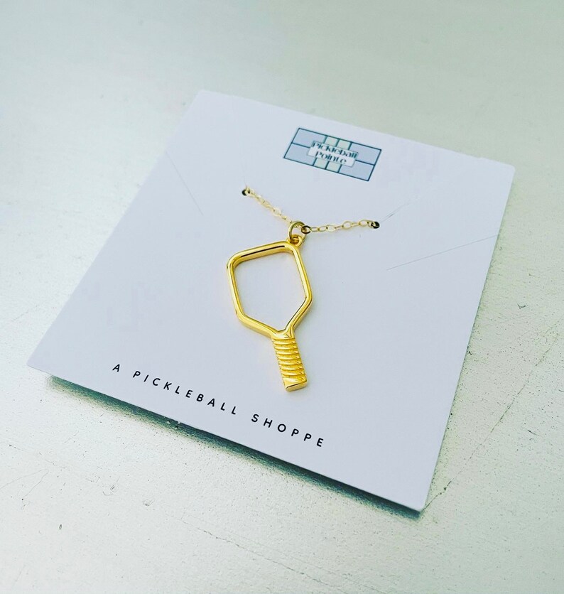 Peut inclure: Un collier en or avec un pendentif en forme de raquette de pickleball. Le pendentif est un simple contour d'une raquette avec un manche textur&eacute;. Le collier est pr&eacute;sent&eacute; sur une carte blanche avec le texte "A Pickleball Shoppe".