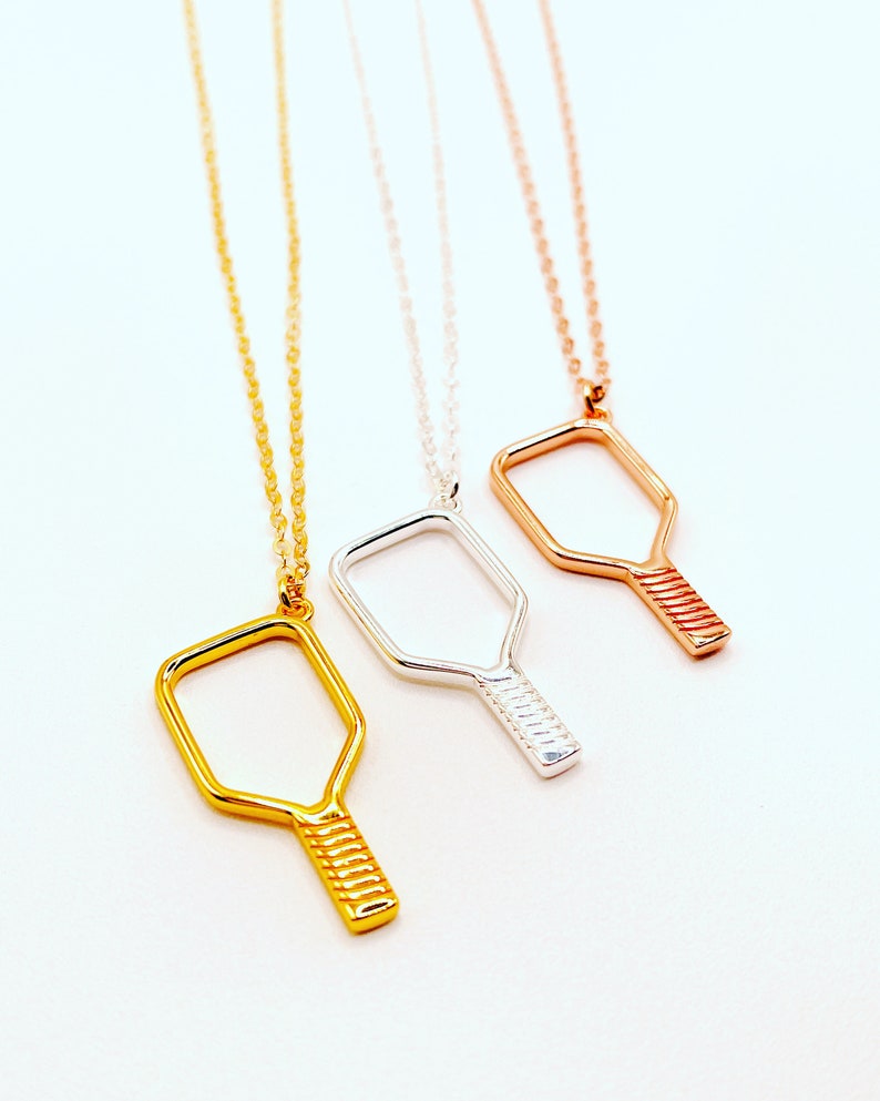 Peut inclure: Trois colliers en argent avec un pendentif en forme de raquette de pickleball. Les colliers sont en or, argent et or rose.