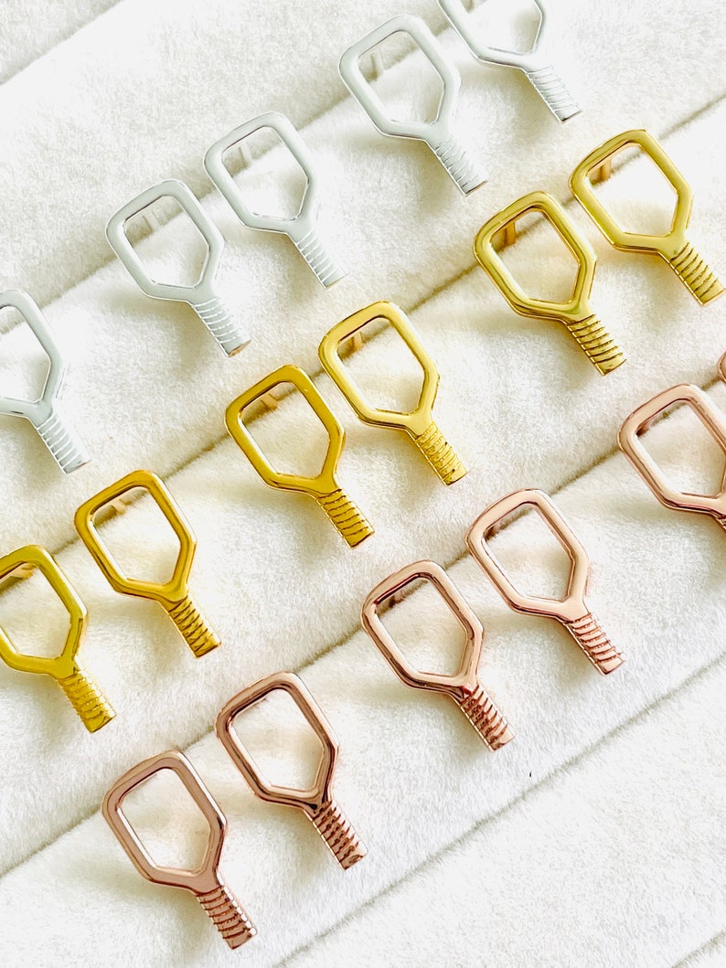 May include: A set of 12 key-shaped earring findings in three different finishes: silver, gold, and rose gold. The findings are arranged in rows on a white velvet background.