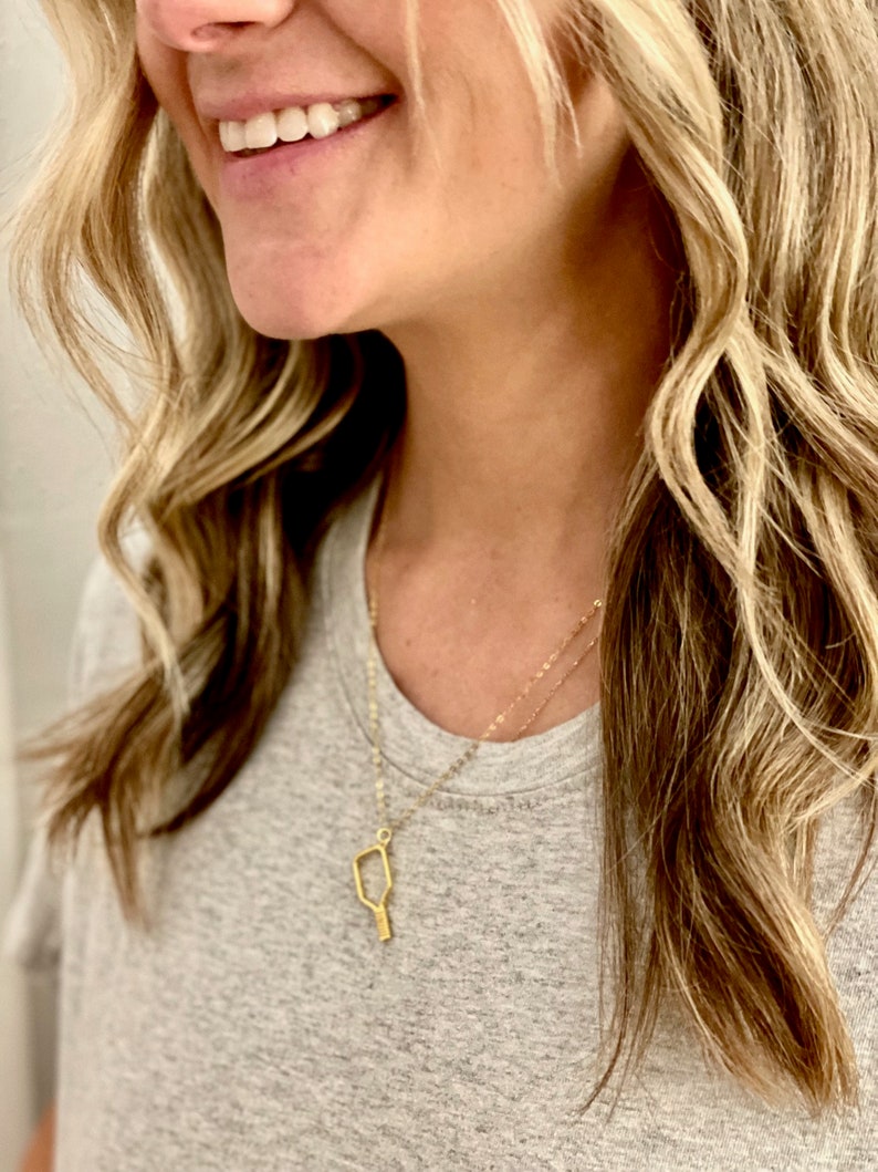 Peut inclure: Un collier en or avec un pendentif en forme de raquette, un &eacute;quipement de sport populaire. Le collier est port&eacute; par une personne aux cheveux longs et blonds.