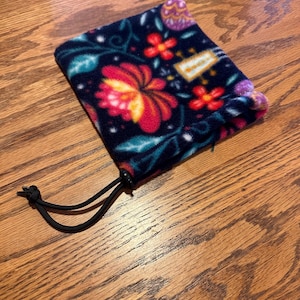 May include: A dark blue fleece pouch with a floral design in red, yellow, and teal. The pouch has a drawstring closure and a small tag with text. The floral pattern includes stylized flowers and leaves.