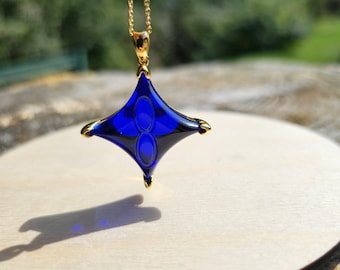May include: A gold chain necklace featuring a blue star-shaped pendant with gold detailing. The pendant's design is symmetrical. The necklace is presented on a light wooden surface, with a blurred green backdrop.