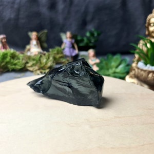 Peut inclure: Un cristal d'obsidienne gris foncé avec une texture rugueuse et irrégulière. Le cristal est posé sur une surface en bois brun clair.