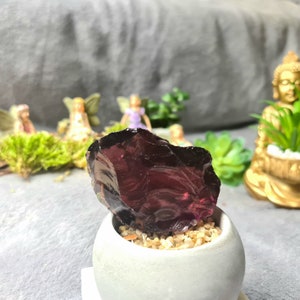 May include: A dark purple obsidian crystal sits in a small white planter with sand. The crystal is smooth and has a reflective surface.