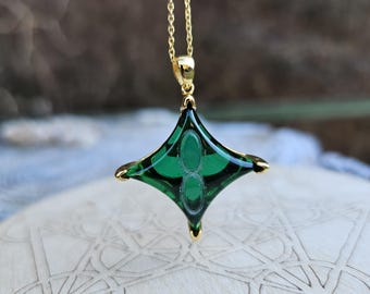 May include: A gold chain necklace with a green glass pendant shaped like a four-pointed star. The pendant has a detailed design with a pattern of circles and lines.