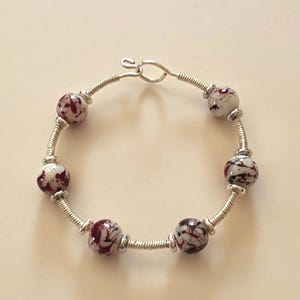 Könnte beinhalten: Ein kreisförmiges Armband mit fünf runden weißen Perlen, die mit dunkelroten und schwarzen Spritzmustern versehen sind. Die Perlen sind durch Segmente aus gewickeltem silberfarbenem Draht und kleinen silberfarbenen Zwischenperlen getrennt.