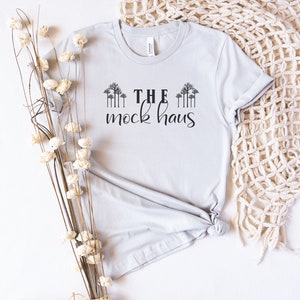 May include: A light gray t-shirt with the text "THE mock haus" in black. The shirt has a rolled sleeve and is knotted at the bottom. Decorative branches with white flowers are on the left side of the shirt.