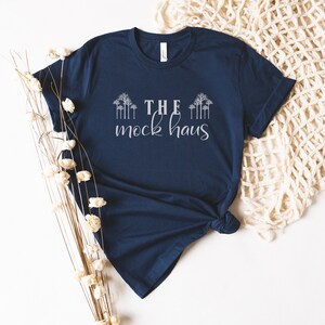 May include: A navy blue t-shirt with the text "THE mock haus" in white lettering. The shirt is laid flat on a white surface with a white crocheted blanket and dried flowers.