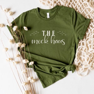 May include: A green t-shirt with the text "THE mock haus" in white lettering. The shirt is folded and laid on a white surface with dried flowers and a white crocheted blanket.