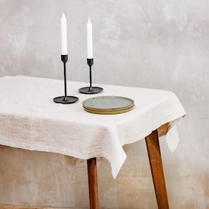 May include: Two black candle holders with white candles on a white linen tablecloth on a wooden table with wooden legs.